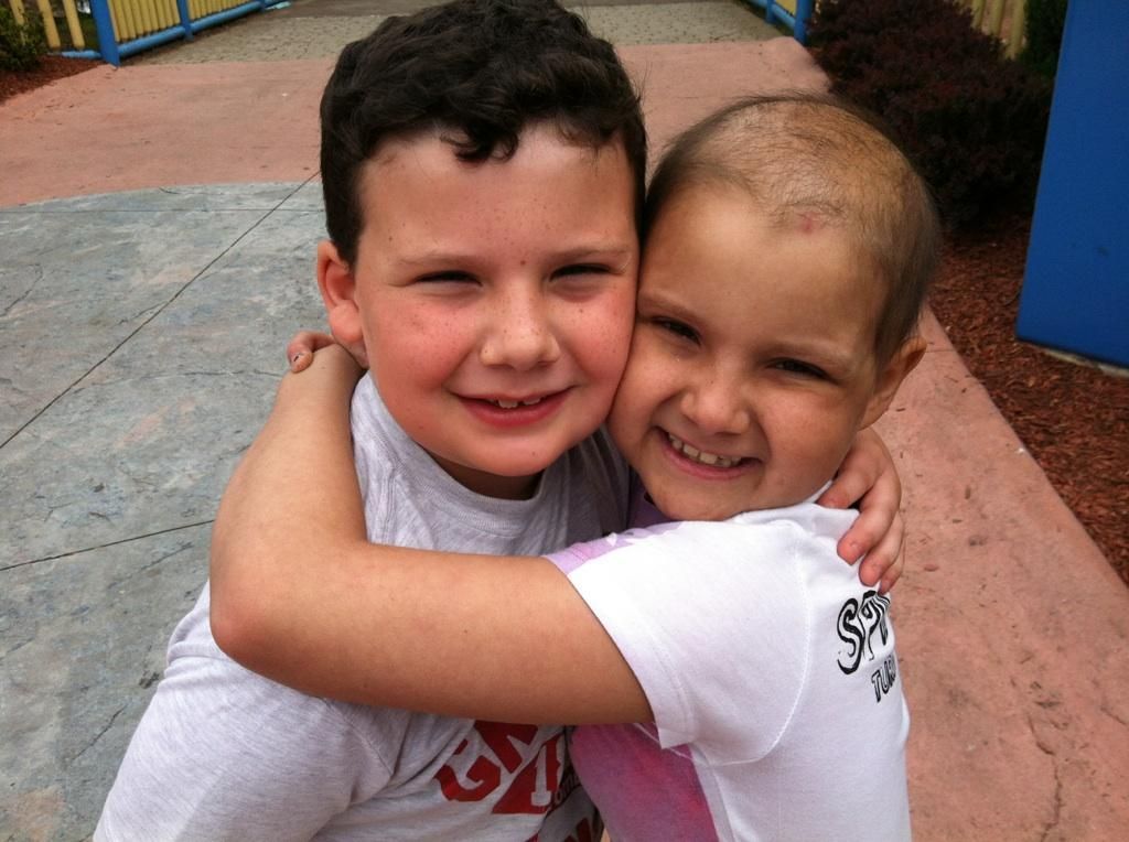 Two smiling children embrace outdoors; one with short hair, the other with a gray shirt.