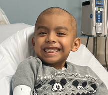 Boy with a shaved head smiles, sitting in a hospital bed with an IV drip and Mickey Mouse sweater.