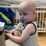 Baby with a bald head, sucking on pacifier, holding a stuffed animal, in a hospital bed.