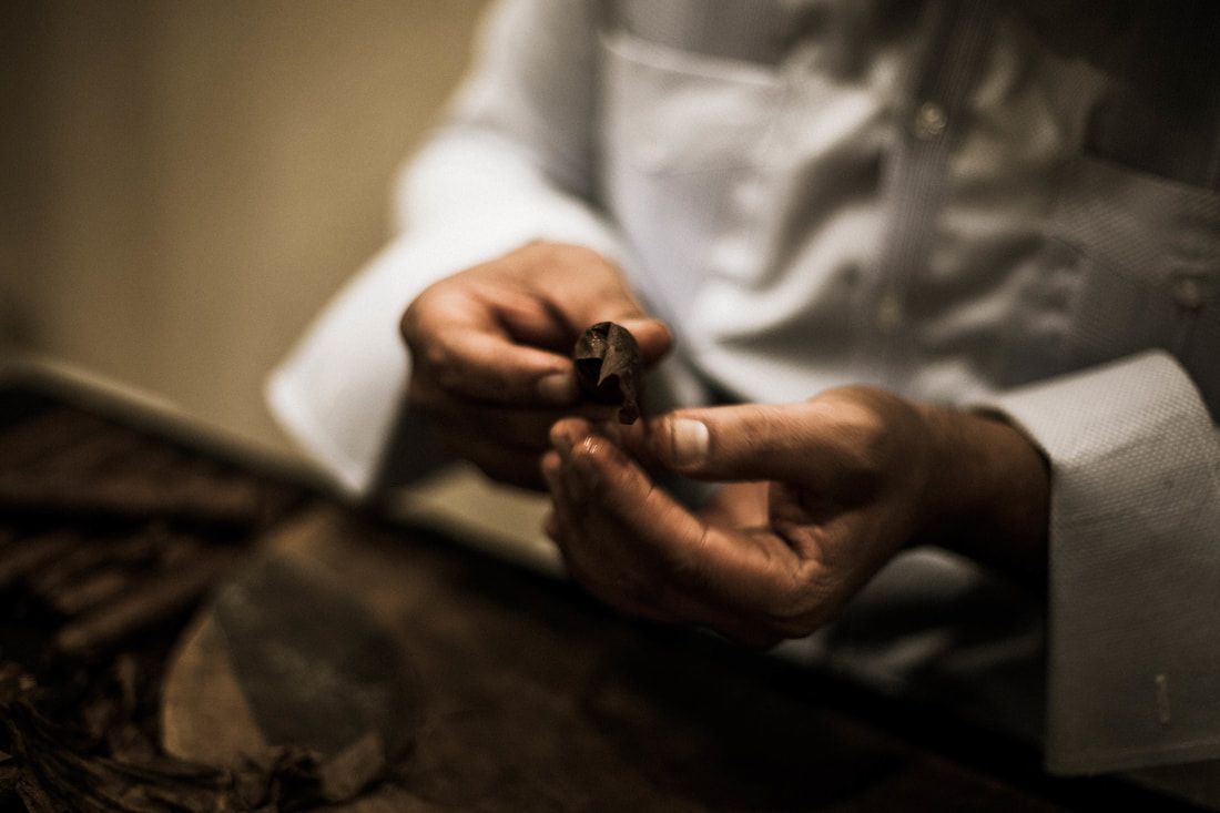 Person's hands holding and examining a small, dark object, possibly tobacco. They wear a white button-down shirt.