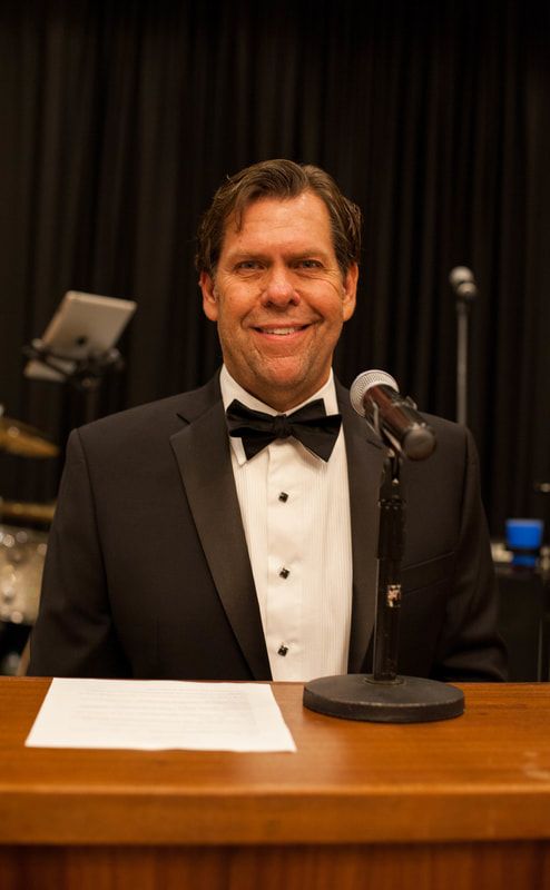Man in tuxedo at podium with microphone, smiling at the camera. Black backdrop, band in background.