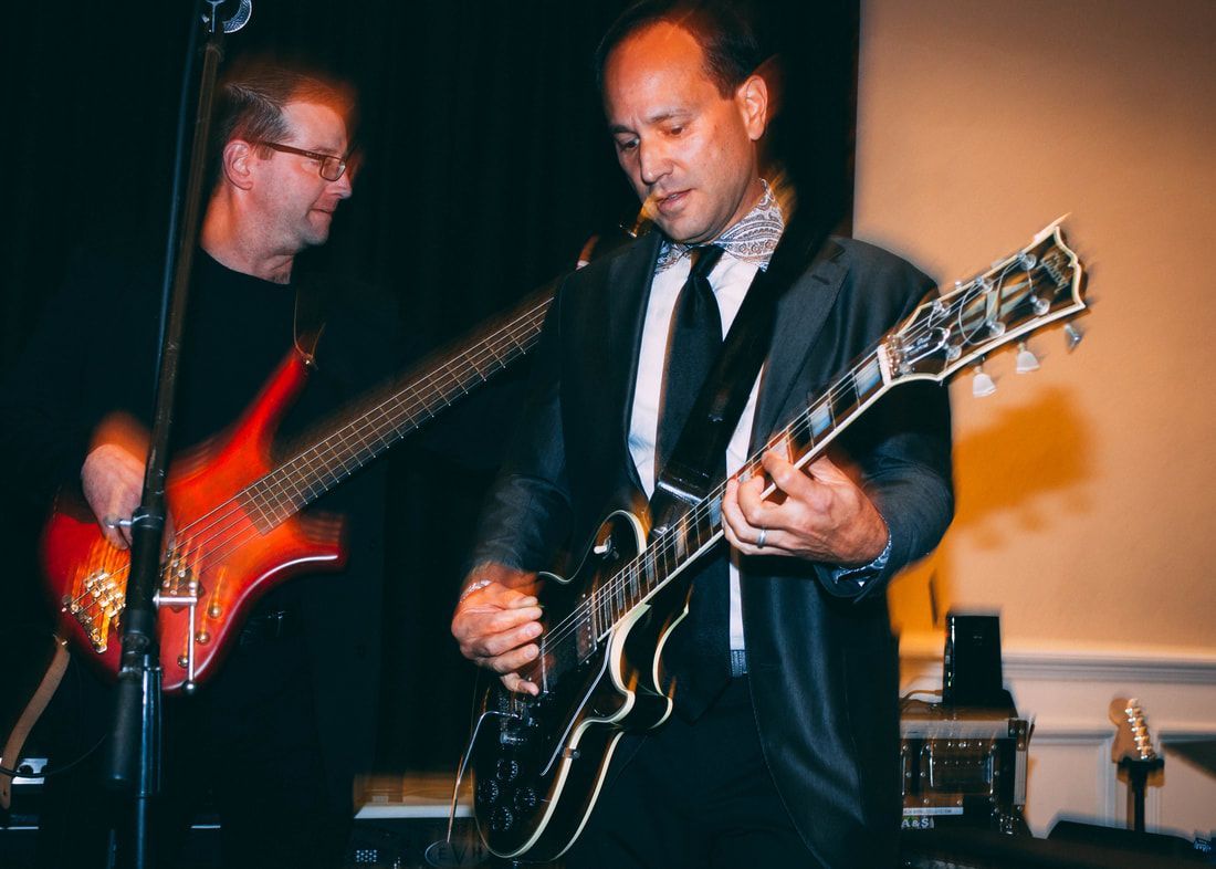Guitarist in suit plays electric guitar as bassist stands behind him on stage.