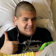Person in a hospital bed gives a thumbs-up. They wear a black shirt and earbuds, smiling.