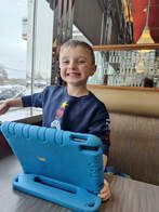 A smiling child sits by a window with a blue tablet in a restaurant.