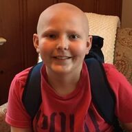 Boy with a shaved head and backpack, smiling, wearing a red shirt, seated indoors.