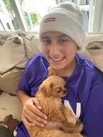 Person in purple shirt and white beanie smiles, holding a small brown puppy.