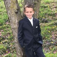 Boy in a dark suit leans against a tree, smiling outdoors.