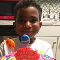 Boy smiling, holding a birthday cake with candles; kitchen in background.
