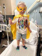Young child in a superhero costume gives a thumbs-up in a hospital room.