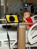 Young child in red shirt sits on a hospital bed watching a tablet on a tray table.