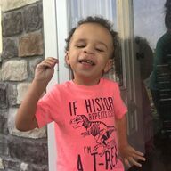 Boy in a pink shirt with a dinosaur graphic smiles outdoors, gesturing with his hand.