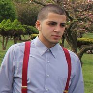 Man in light blue shirt with red suspenders, standing outdoors near trees and bushes.