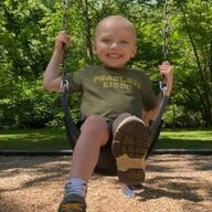 Boy on a swing smiles at the camera, wearing green shirt and khaki shoes, in a park.