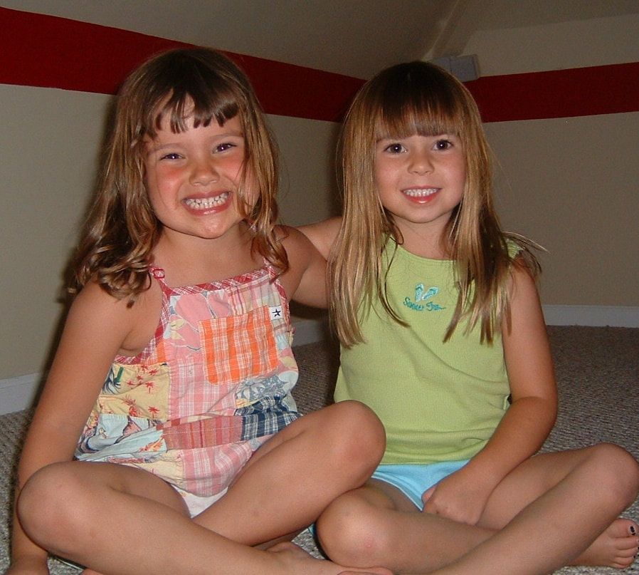 Two young girls smile, sitting cross-legged indoors. One wears a floral tank top, the other a green shirt.