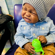A young child wearing a gray beanie and denim shirt cries while holding a green sippy cup.