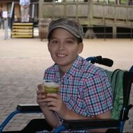 Person in wheelchair holding a drink, smiling, wearing a plaid shirt and cap. Outdoors, in front of a structure.