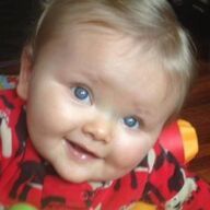 Baby with light blonde hair and bright blue eyes smiles, wearing a red patterned onesie.