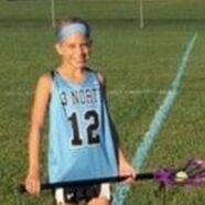 Girl in a light blue lacrosse jersey with number 12, holding a lacrosse stick on a field.