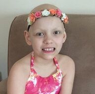 Girl with a bald head wearing a floral headband and halter top, smiling. Sitting indoors.