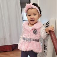 Child in a pink fluffy vest, headband, and gray belt, smiling. Leaning on a railing near a white vent.