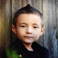 Boy with short, spiky hair and dark eyes, wearing a black top, leaning against a wooden fence.