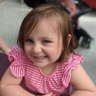Girl with brown hair and a pink striped shirt smiles, outside.