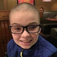 Young person with shaved head and glasses smiles in a restaurant booth, wearing a blue jacket.