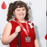 Young person in red dress holds a cane, wearing a medal, smiling at a red carpet event.