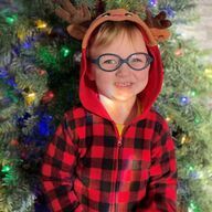 Child in reindeer antlers and red plaid onesie smiles in front of Christmas tree.