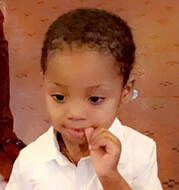 Child with brown eyes and short hair biting a finger. Wearing a white collared shirt.