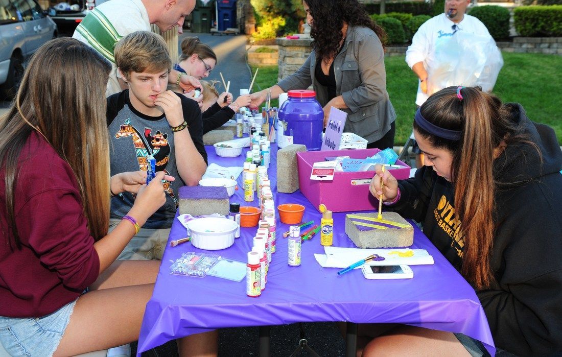People painting at a table outdoors; purple tablecloth, paint, brushes, and smiling faces.