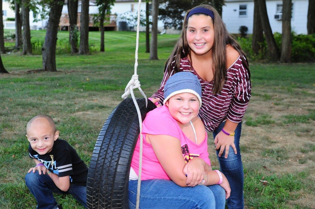 Three children with a tire swing in a grassy yard. Smiling, one sits on the tire, one crouches, one stands.