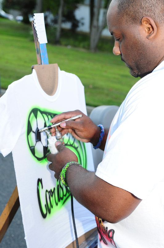 Man airbrushes a soccer design onto a white t-shirt on an easel outdoors. Green, black, and white colors are used.