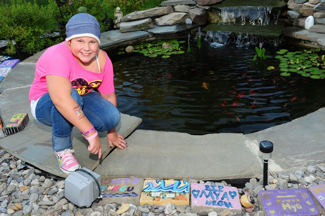 Girl in pink shirt and jeans near a pond, smiling. Stone border, colorful tiles, water feature.