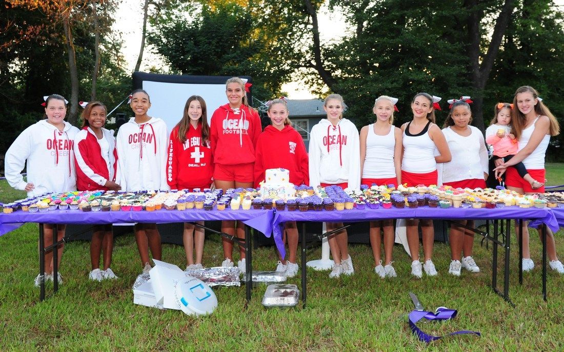 Group of people in red and white clothing behind a table of cupcakes, outdoors.