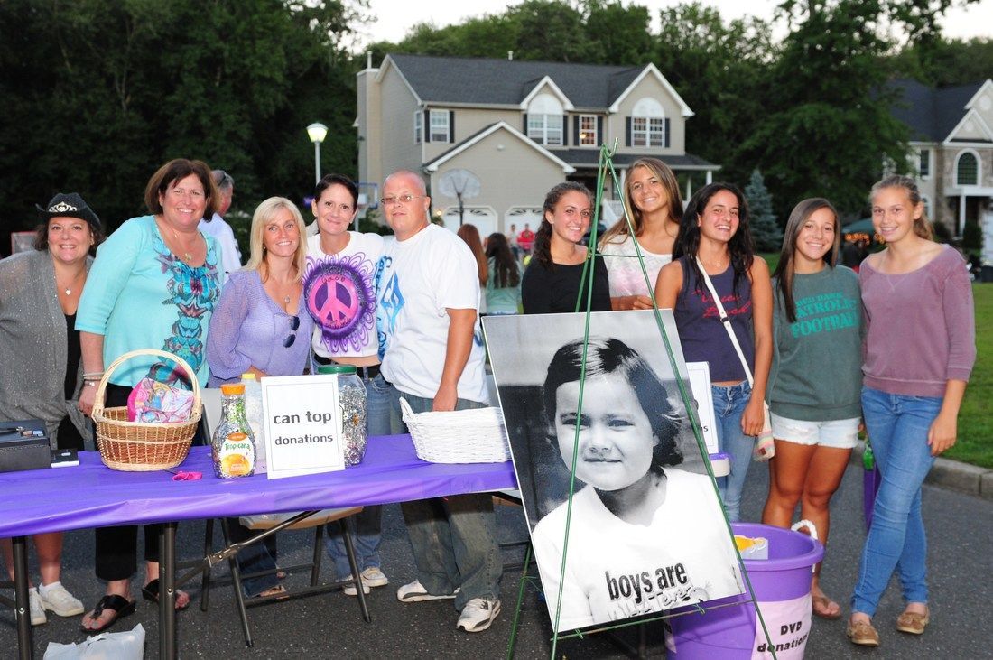 People at table with photo, fundraising event. Purple table, sign, basket. Houses in background.