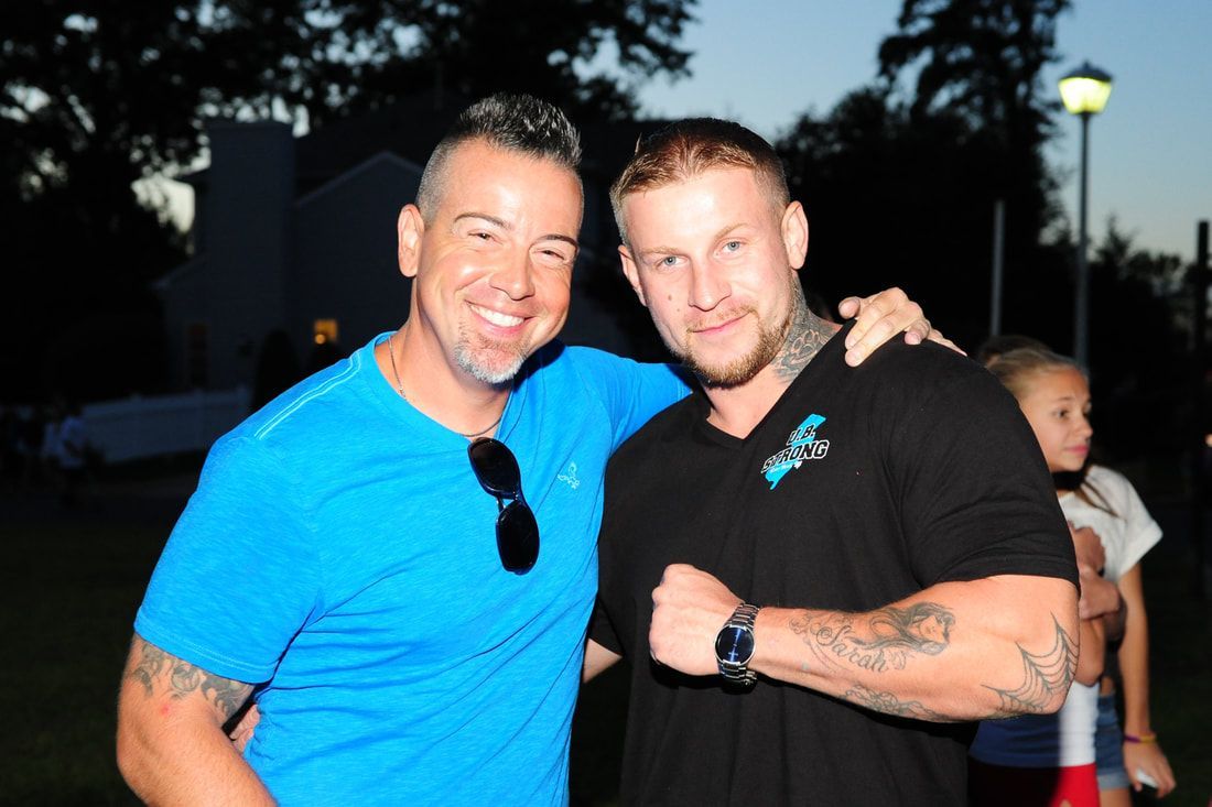 Two men smiling outdoors with one arm around the other; one in blue, one in black; dusk.