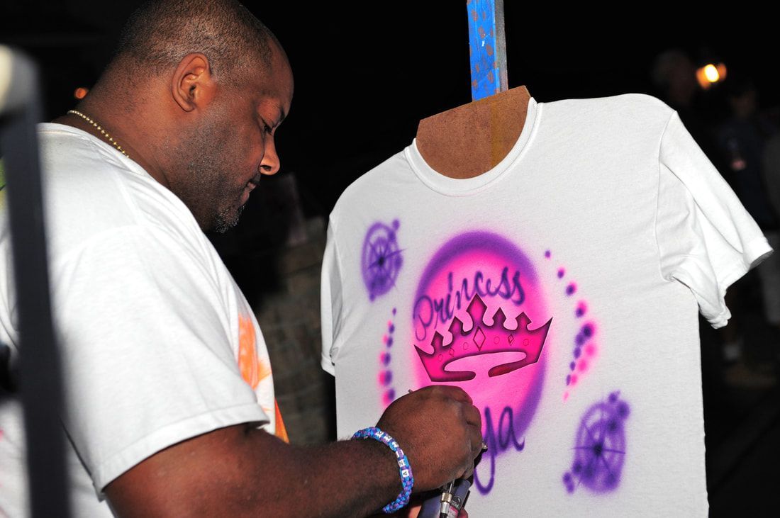 Man airbrushes a white t-shirt with a pink and purple crown design. Night setting.