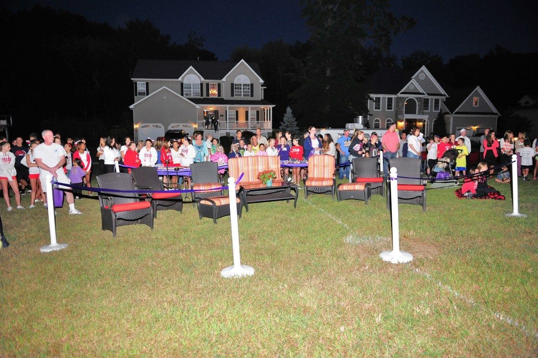 People gathered at night outdoors. Lawn chairs face a crowd, houses in background.