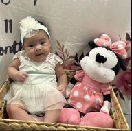 Baby in a light blue dress and white headband sits next to a Minnie Mouse plush in a wicker basket.