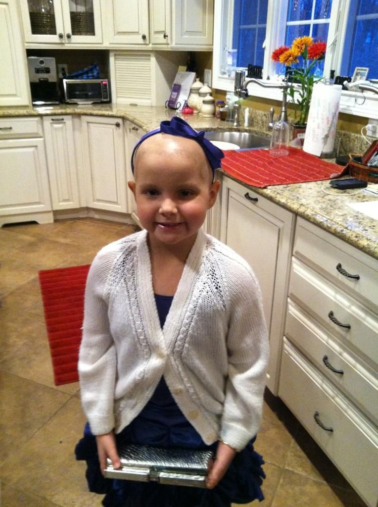 Girl with shaved head and blue bow smiles, holds silver clutch, wearing white cardigan and blue dress in a kitchen.