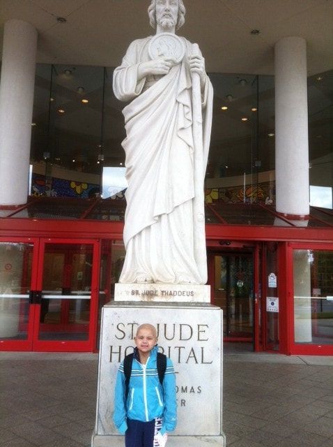 Child in front of St. Jude Hospital, statue of St. Jude. The child has no hair, wearing blue jacket.