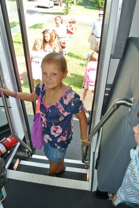 Girl on bus steps, smiling, holding purple bag. Other children and adults outside.