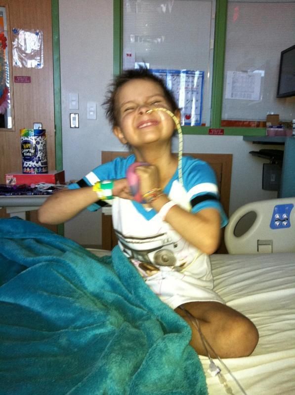 Child in hospital bed smiling, holding small ball, wearing medical tubing.