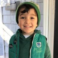 Young child with dark hair, wearing a green hat and vest, smiling outside.
