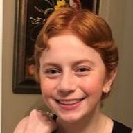 Smiling person with red hair, posing, against a blurred background.