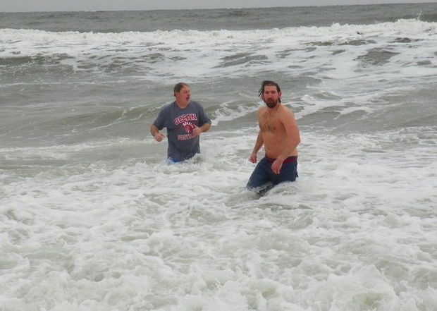Two people wading in ocean waves, one shirtless, both wearing dark pants. Gray, overcast sky.