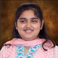 Woman with dark hair, wearing a pink patterned top, smiling. Background is blurred brown.