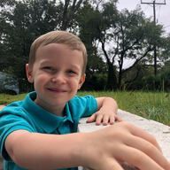 Boy with a small smile, blue shirt, leaning on a concrete surface, outdoors with trees in the background.
