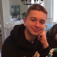 Young person in black hoodie, resting chin on hand, smiling indoors.